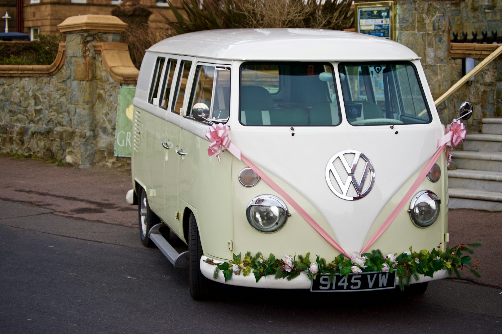 Svadobný Volkswagen Bus v krémovej farbe, ozdobený ružovými stuhami a zeleňou.